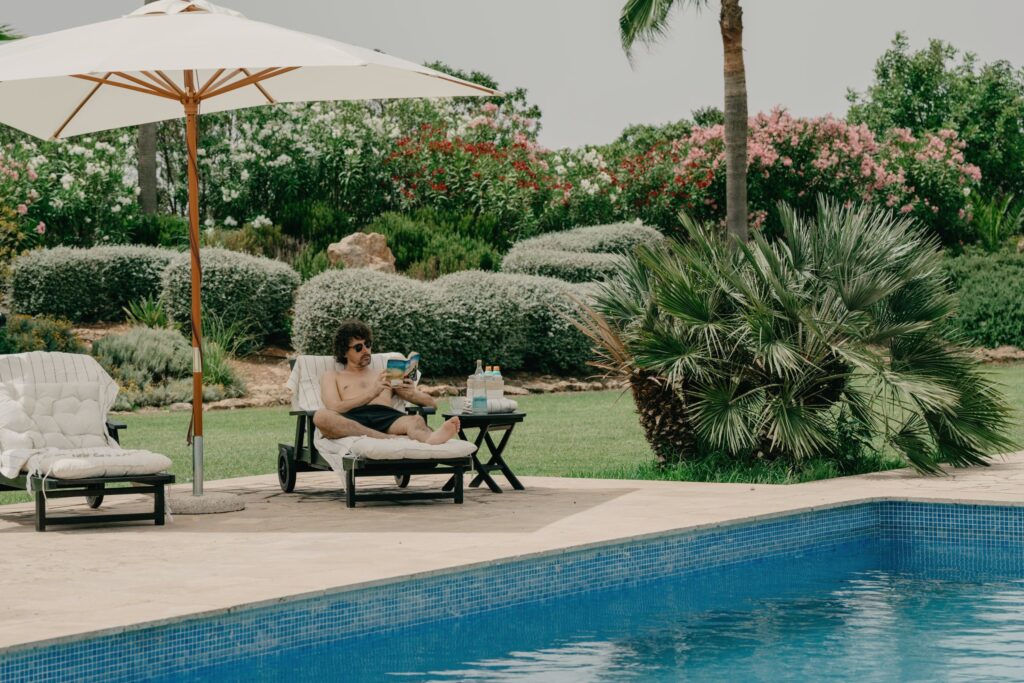 Man reading on a sun lounger beside a swimming pool under a white umbrella