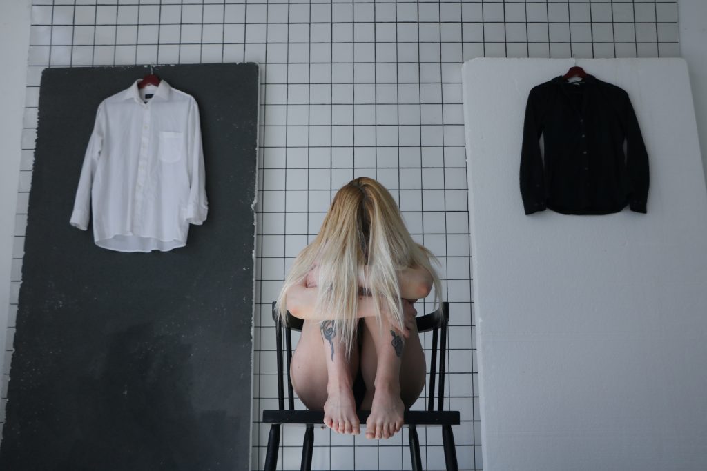 A girl sitting on a chair while hugging her legs to her chest, two shirts are hung behind her.