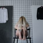 A girl sitting on a chair while hugging her legs to her chest, two shirts are hung behind her.