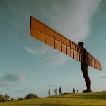 A big statue of a human with airplane wings as arms with people walking beneath.