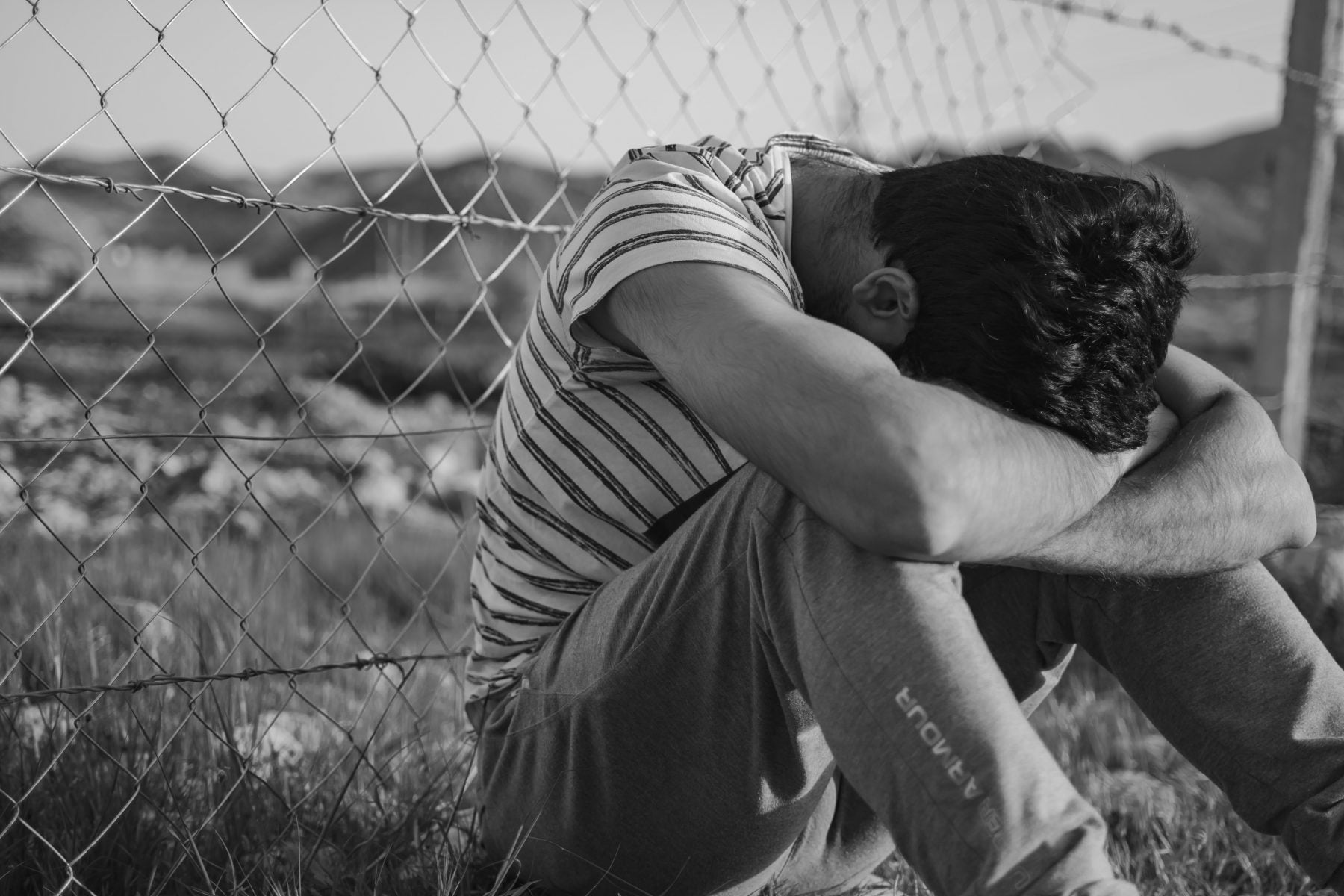 Black and white image of a person sitting on the ground with his head between his hands.