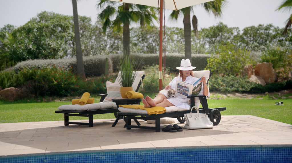 Woman relaxes on a poolside lounger reading a magazine under an umbrella, with a small dog on her lap
